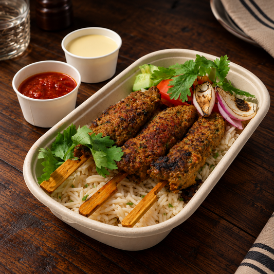A prepared food dish in a beige paper tray on a dark wooden table, featuring grilled meat skewers over rice with garnish of cilantro, tomato, onion, and side cups of sauce.